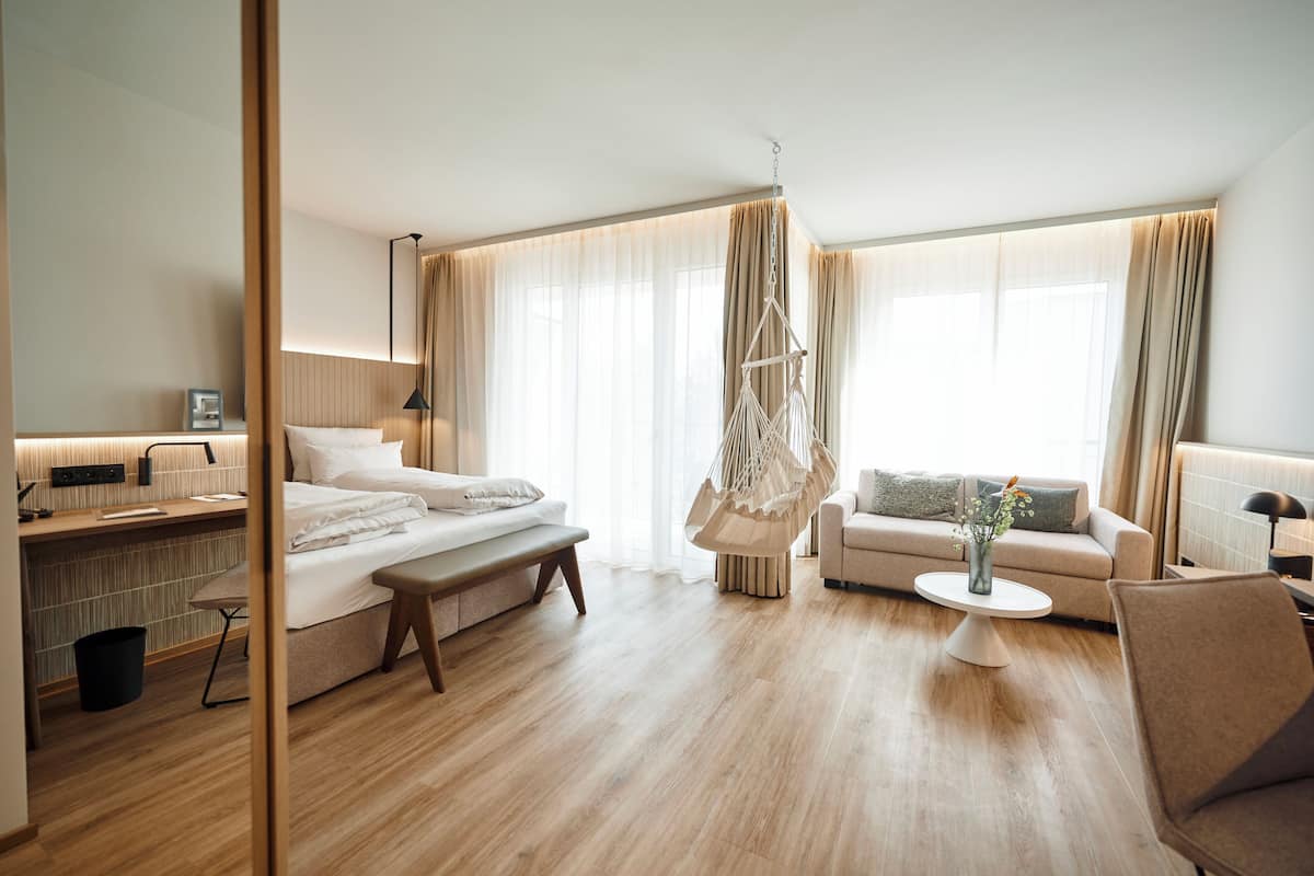 A modern hotel room with a large bed, a sofa, a hanging chair, a small table with a vase, and a desk with a lamp.