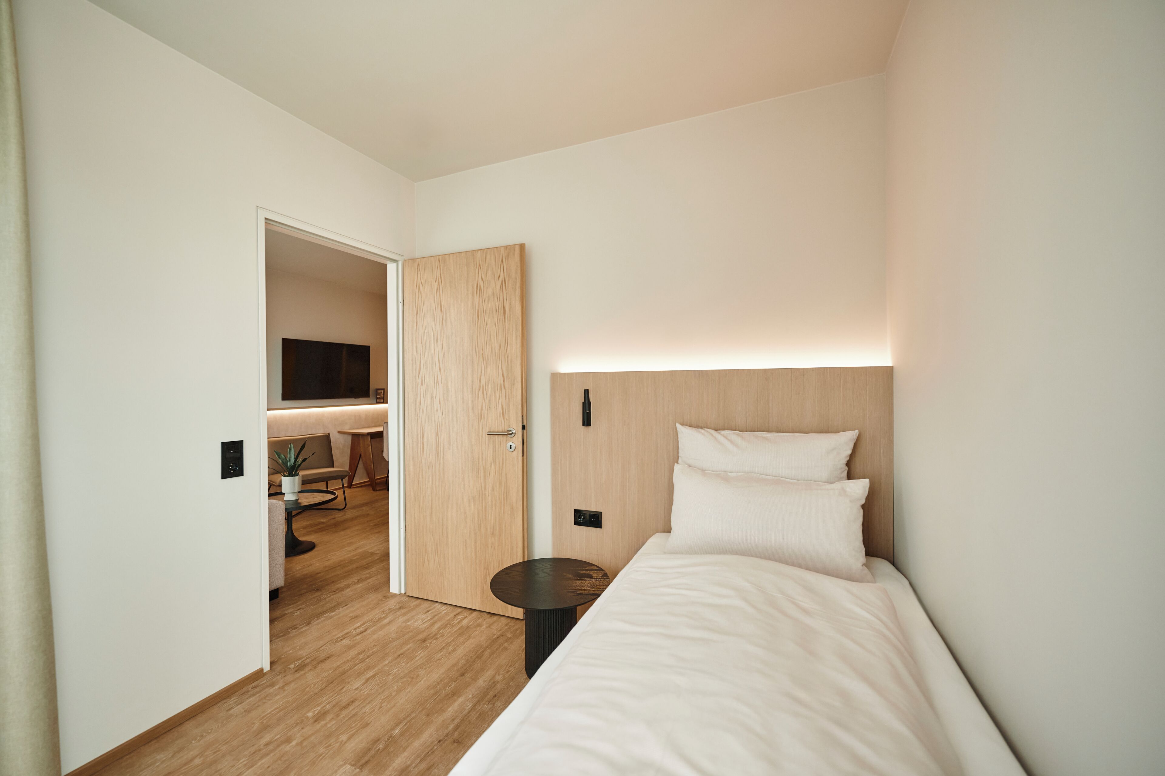 Quarto de hotel moderno com piso de madeira, cama com roupa de cama branca, mesa lateral redonda e um pequeno vaso de planta.