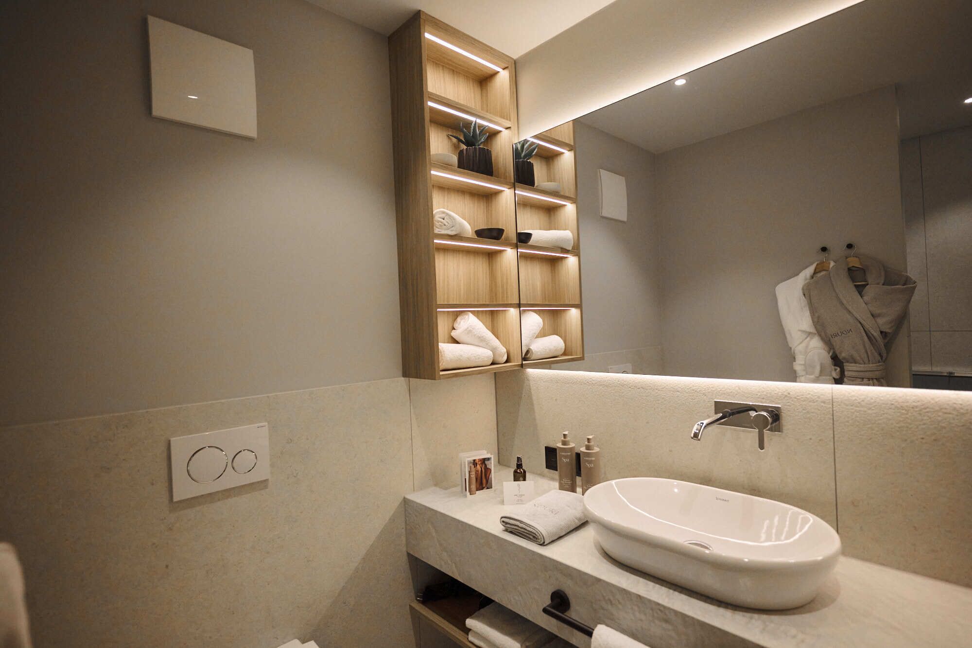 A modern bathroom with a large mirror, a built-in wooden shelf with towels, a white sink, and a towel rack.