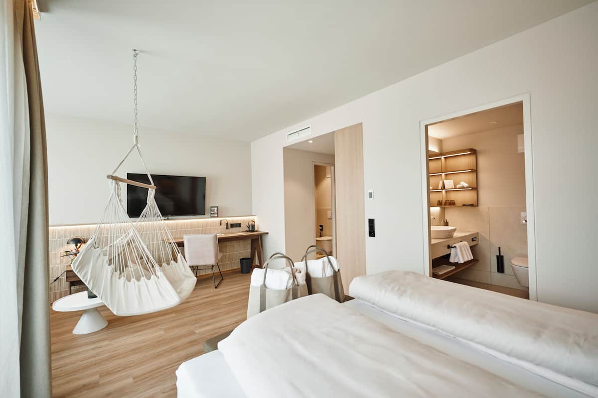 A modern hotel room with a large bed, a hanging chair, a desk with a chair, and a bathroom visible through an open door.