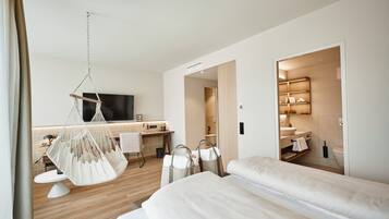 A modern hotel room with a large bed, a hanging chair, a desk with a chair, and a bathroom visible through an open door.