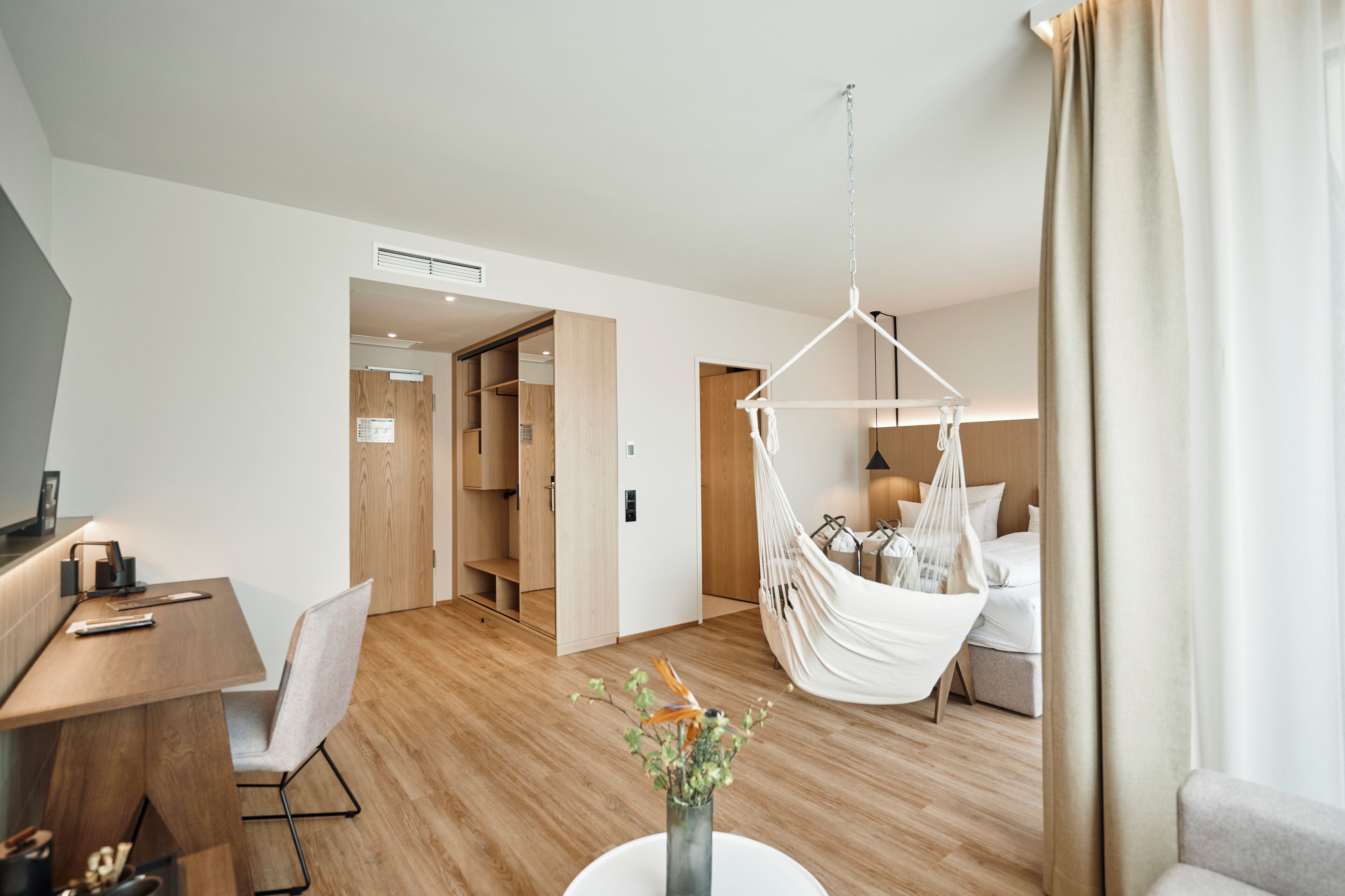 Une chambre d’hôtel moderne avec un plancher en bois, un bureau avec une chaise, un fauteuil suspendu, un lit et une table de chevet.