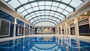 Una amplia piscina interior con techo de vidrio, rodeada de paredes con azulejos azules y un mural.