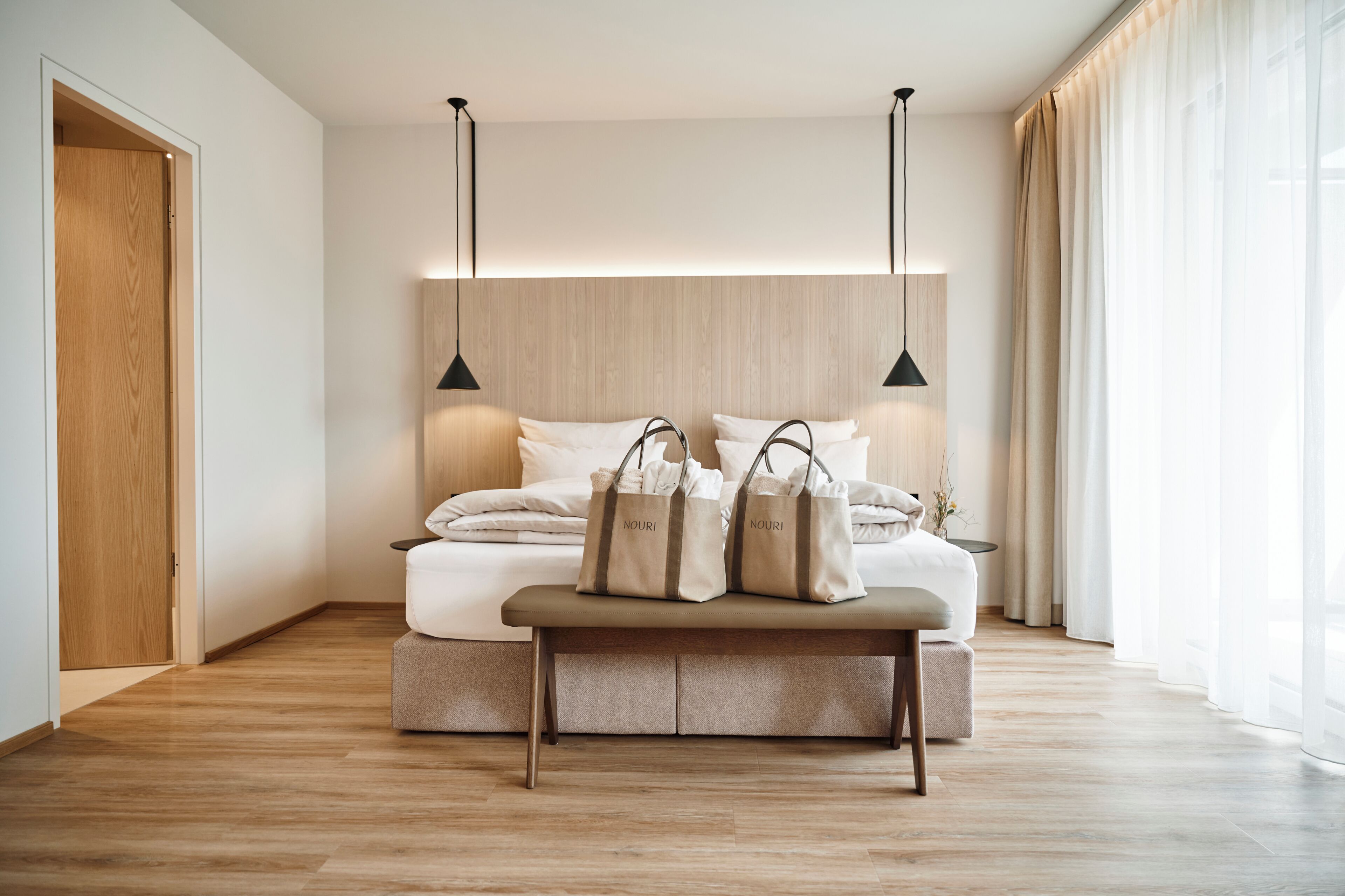 A modern bedroom with a wooden floor, a bed with white linens, two pendant lights, a bench with bags, and a large window with curtains.