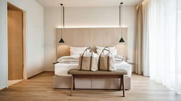 A modern bedroom with a wooden floor, a bed with white linens, two pendant lights, a bench with bags, and a large window with curtains.
