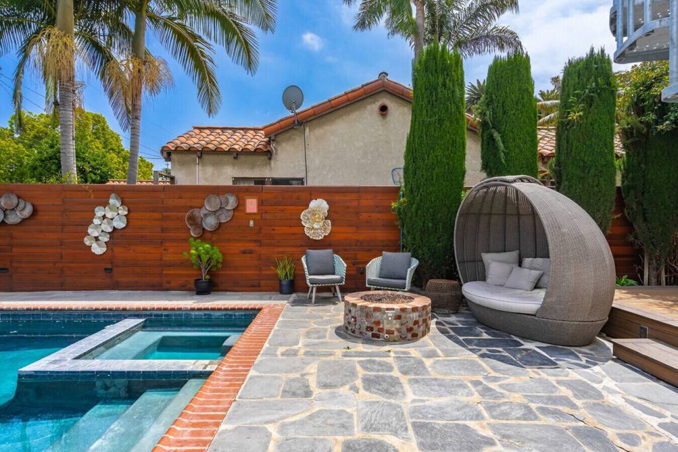 A swimming pool with stone tiles, a wooden wall with decorative shells, and a round seating area with cushions.