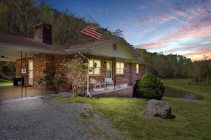 A brick house with a porch, an American flag, and a pond in the background.