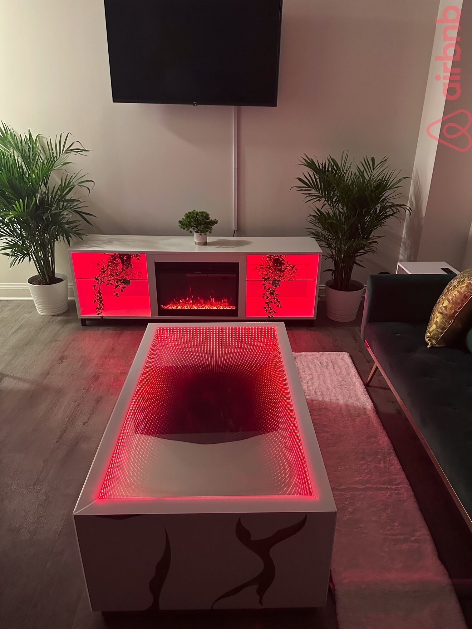 A modern living room with a central heated floor, a flat-screen TV mounted on the wall, and two potted plants on either side of a sofa.