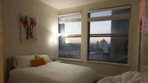 A hotel room with a large bed, a white headboard, a painting of an elephant, and a view of a cityscape through the windows. - Luxurious Downtown Indy City View Apartment!  (Indianapolis)