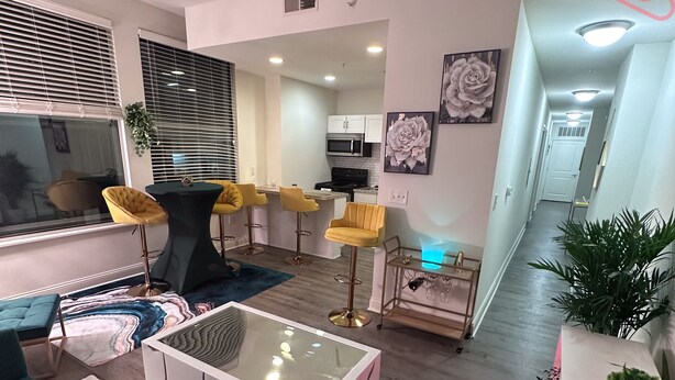 A modern living room with a sofa, a coffee table, and yellow bar stools. There is a kitchen area in the background with a microwave and cabinets. - Luxurious Downtown Indy City View Apartment!  (Indianapolis)