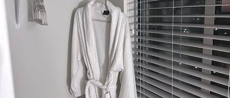Ein Badezimmer mit einem weißen Bademantel, der an einem Haken hängt, einem Handtuchhalter mit gefalteten Handtüchern und einem Fenster mit Jalousien.