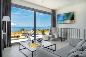 Un salon moderne doté d’une grande porte-fenêtre coulissante donnant sur un balcon avec vue sur la mer. On y trouve un canapé, un fauteuil et une table basse agrémentée de livres et d’objets décoratifs.