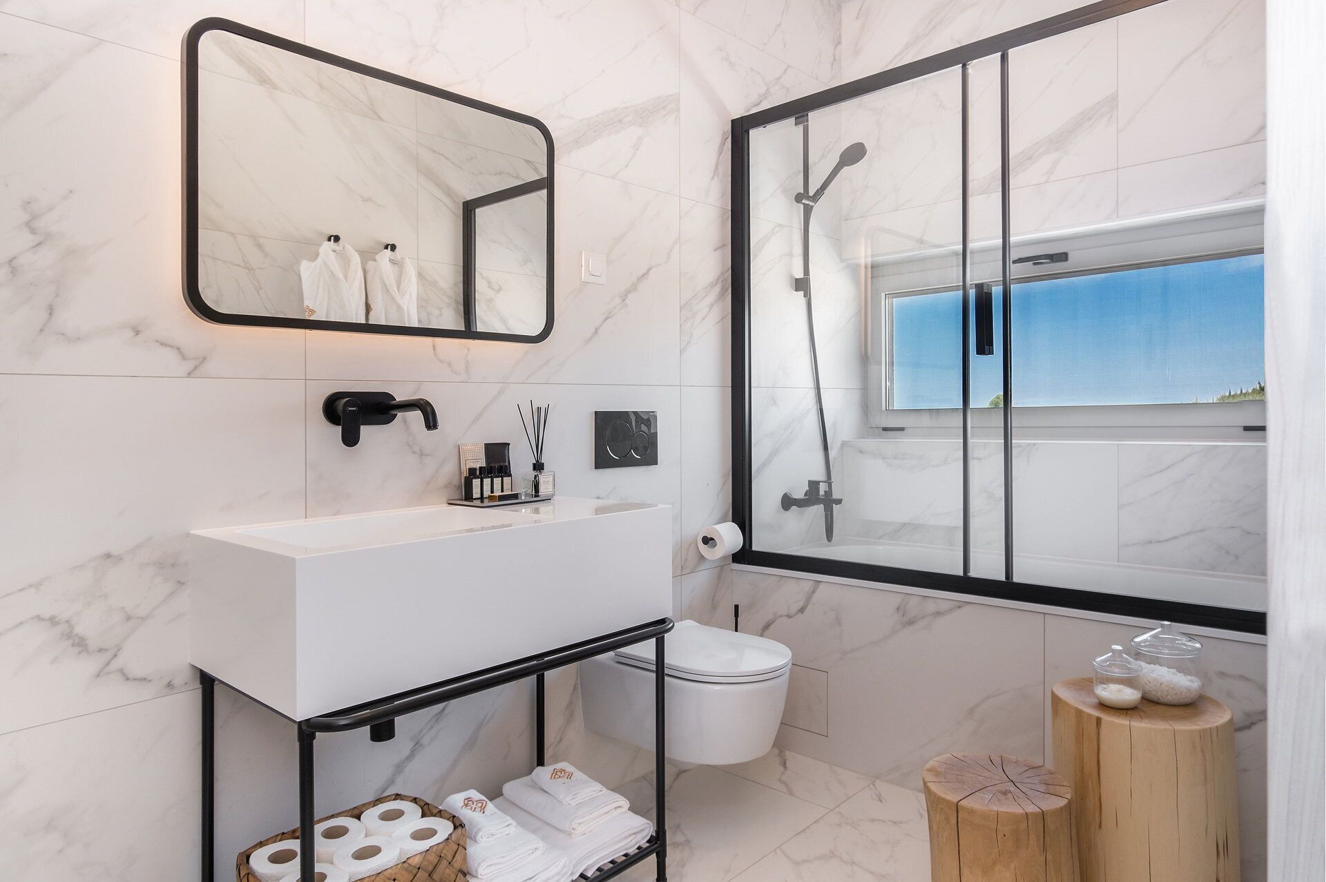 A modern bathroom with a white marble sink, a black faucet, a glass shower enclosure, and a wooden stool with toiletries.