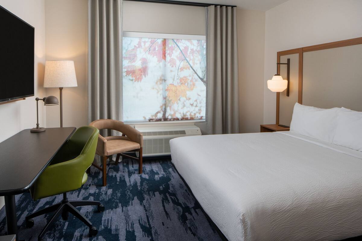Une chambre d’hôtel comprenant un lit, un bureau avec une chaise, une télévision, une fenêtre avec des rideaux et une lampe.
