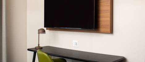 A modern hotel room with a flat-screen TV mounted on the wall, a small desk with a chair, and a lamp.