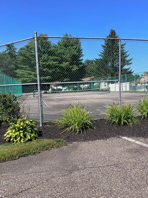 Ein von einer Hecke umgebener Tennisplatz mit Parkplatz und einem Gebäude im Hintergrund.