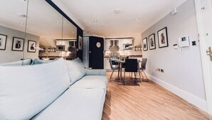 A modern living room with a sofa, a dining area with a table and chairs, and a kitchen in the background. - The Apartment, Broadway (Broadway)