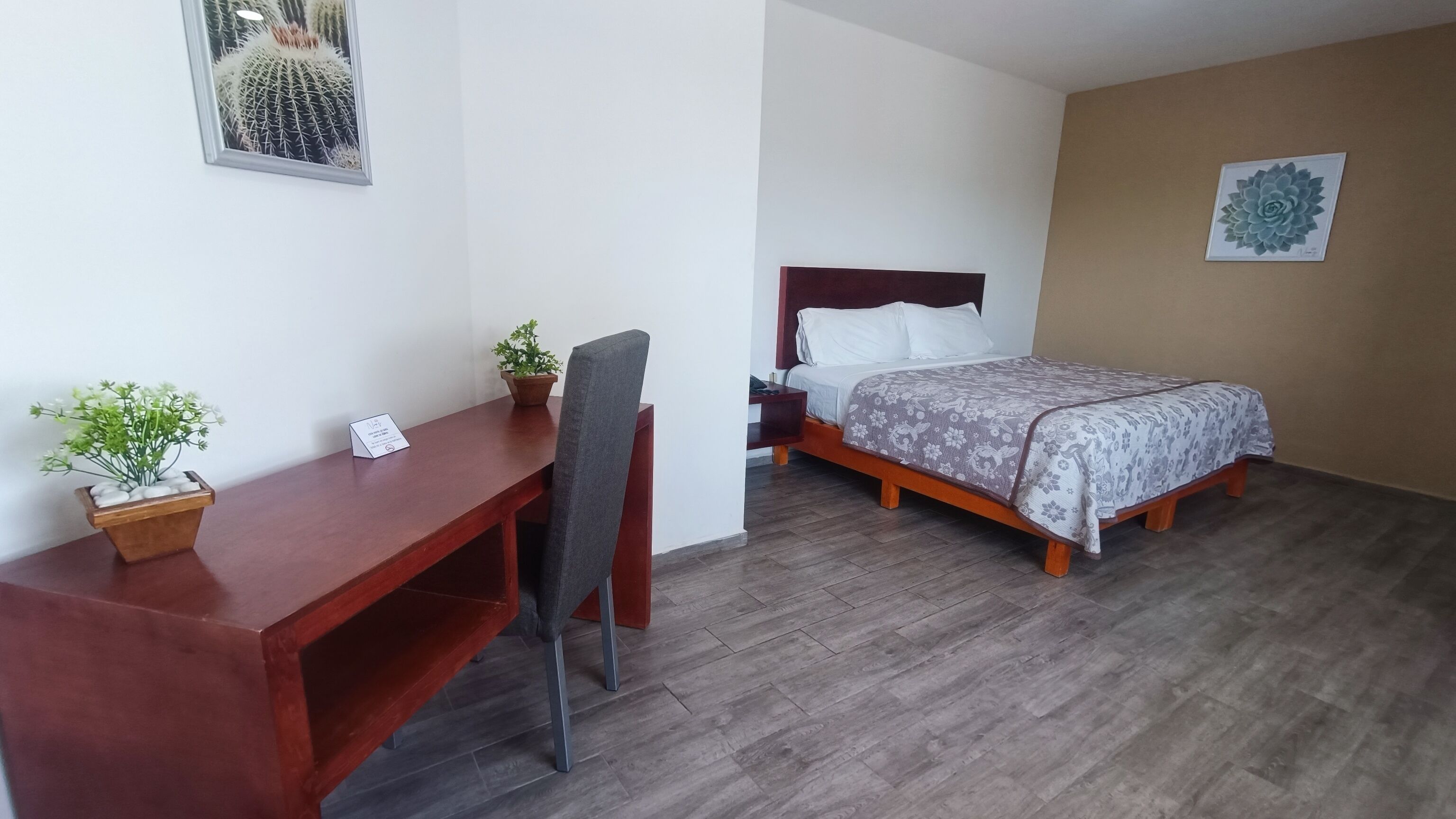 Quarto de hotel com escrivaninha de madeira, uma cadeira, cama com colcha florida, mesa de cabeceira, planta em vaso e um quadro na parede.