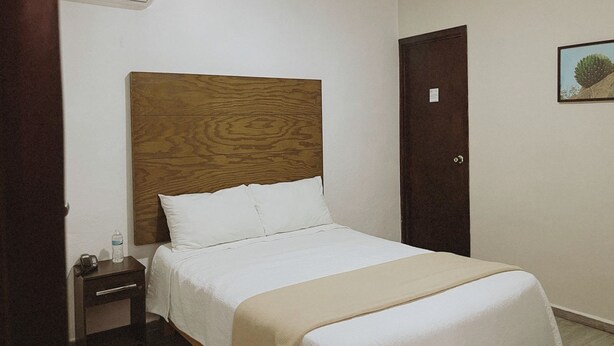 A hotel room with a wooden headboard, a single bed with white bedding, a nightstand with a water bottle and phone, and a door with a small picture on the wall. - Noas hotel (Matamoros)