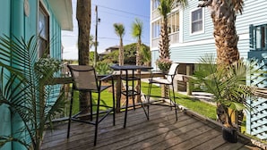 Una terraza de madera con mobiliario exterior, rodeada de plantas tropicales y una casa azul.