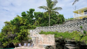 Un escalier en pierre mène à un balcon donnant sur une zone aménagée autour de la piscine, le tout étant entouré d’une végétation luxuriante et surplombé par un ciel bleu clair.