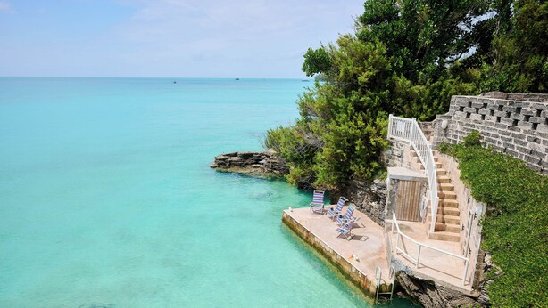 Une vue sur la côte, avec une eau turquoise et limpide, un escalier en pierre menant à une plateforme où se trouvent deux chaises longues, et un promontoire rocheux parsemé d’arbres.