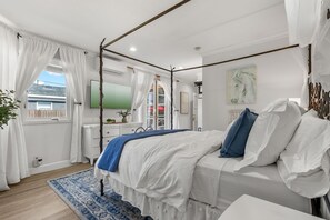 A bedroom with a four-poster bed, a large window with curtains, a bedside table, and a wall-mounted TV.