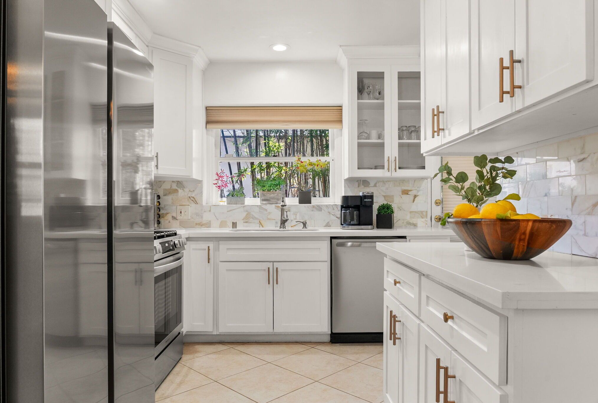 A modern kitchen with white cabinets, stainless steel appliances, a window with a view of greenery, and a bowl of lemons on the counter.