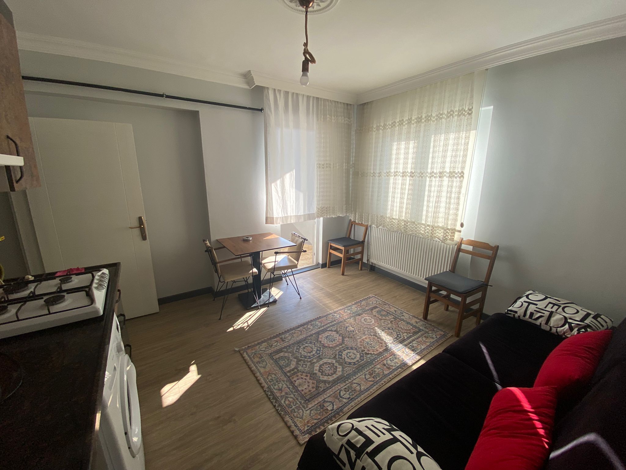 A living room with a sofa, a dining table with chairs, a radiator, and a window with sheer curtains.