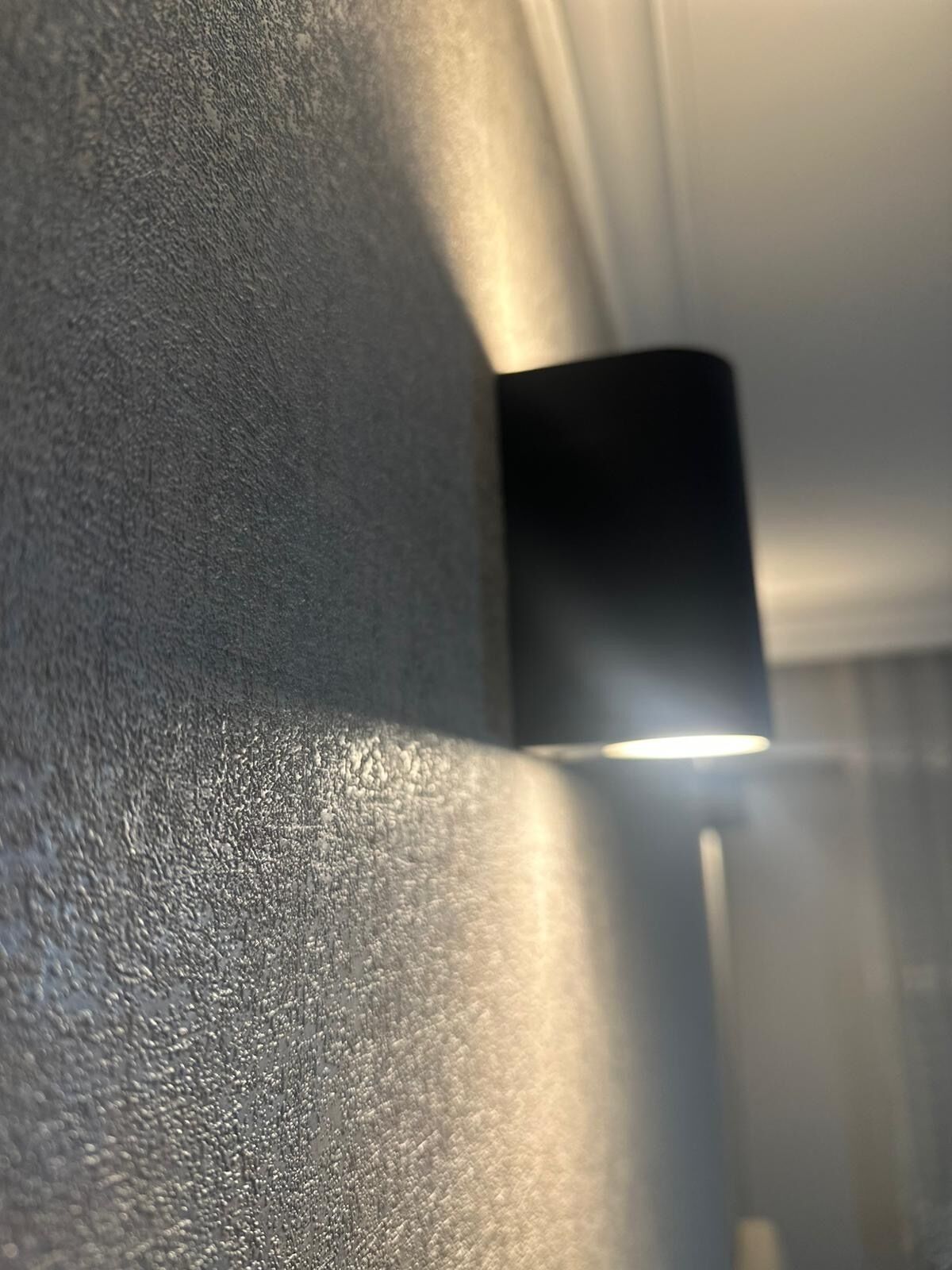 Primer plano de una pared con textura y un objeto negro, posiblemente una lámpara, que proyecta una sombra.