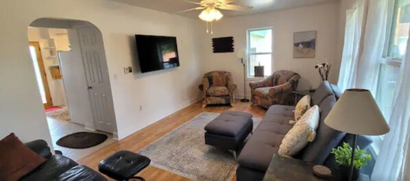 A living room with a sofa, ottoman, chairs, a TV, and a ceiling fan.