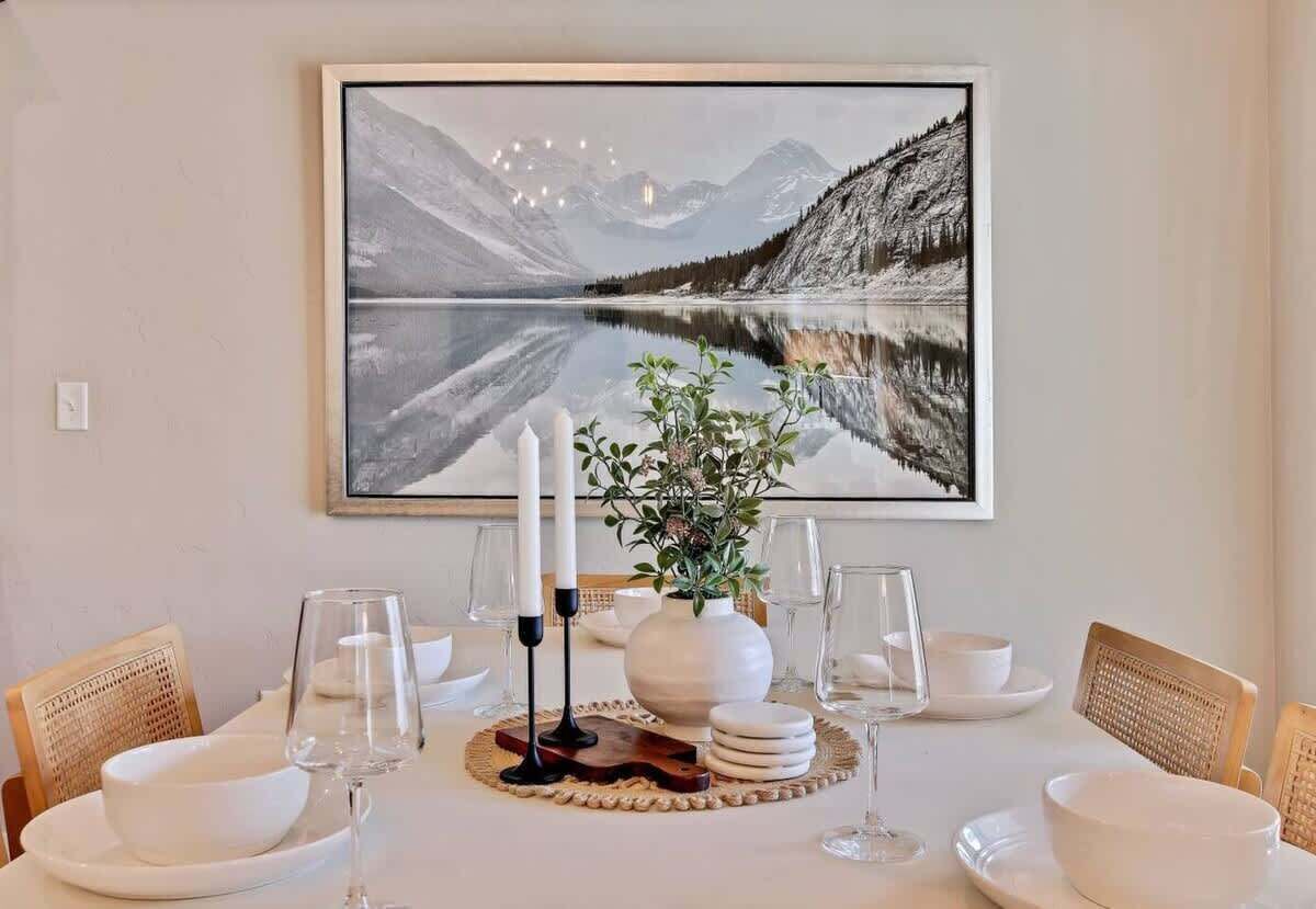 Et spisebord dækket med hvide tallerkner, skåle og glas, en vase med blomster, og et indrammet landskabsmaleri hængende over.