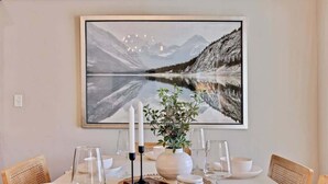 Une table à manger dressée avec de l’assiette, des bols et des verres blancs, un vase avec une plante, et un tableau représentant un paysage, encadré, accroché au-dessus.