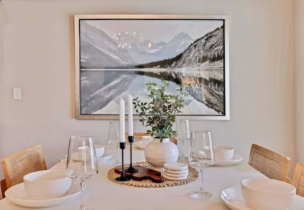 Une table à manger dressée avec de l’assiette, des bols et des verres blancs, un vase avec une plante, et un tableau représentant un paysage, encadré, accroché au-dessus.
