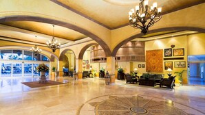 Eine großzügige Lobby mit hohen Decken, Rundbögen und einem zentralen Kronleuchter. Es gibt Sitzbereiche mit Sofas und Topfpflanzen.