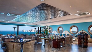 Ein Restaurant mit Korbmöbeln, einem zentralen Tisch mit Blumenvase und Blick auf das Meer.