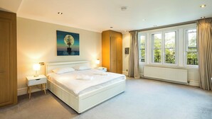 A spacious bedroom with a large bed, a nightstand with a lamp, a wooden wardrobe, and a window with curtains.