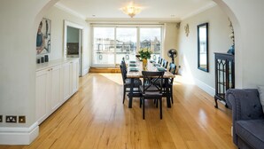 A dining area with a wooden table set for six, surrounded by chairs, with a view of the city through large windows.