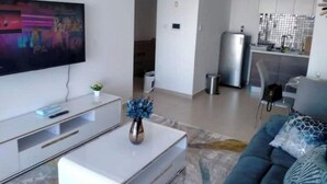 Un salon moderne avec un téléviseur à écran plat, une table basse, un canapé et un coin cuisine à l’arrière-plan.