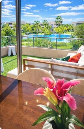 A room with a view of a swimming pool and palm trees, a wooden table, and a chair with a cushion.