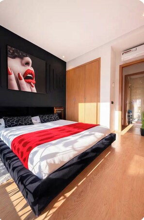 A modern bedroom with a large bed, a black wall with a framed artwork, wooden wardrobes, and a visible bathroom door.