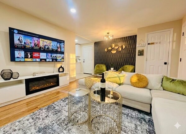 A modern living room with a flat-screen TV, a fireplace, a glass coffee table, and a sofa with cushions.