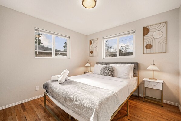 Un dormitorio con una cama, dos mesitas de noche, una lámpara de pared y dos ventanas con persianas.