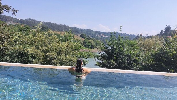 Vista chiara di una piscina con una persona che nuota, circondata da vegetazione lussureggiante e una collinata sullo sfondo.