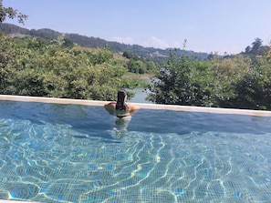 A clear view of a swimming pool with a person in the water, surrounded by lush greenery and a distant view of hills. - Casa da Flor 1 (Vila Caiz - Amarante)
