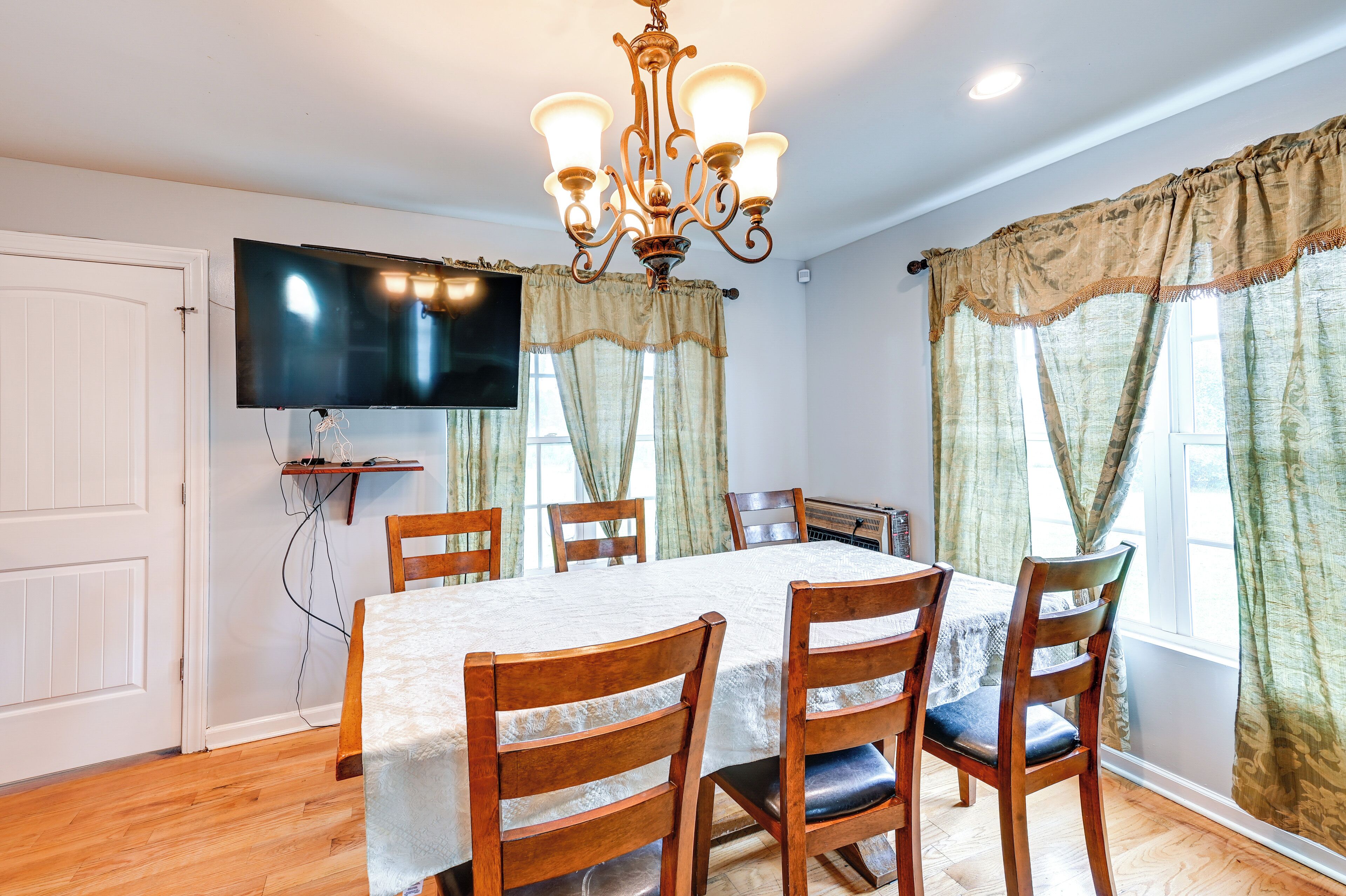 Une salle à manger avec une table dressée pour six personnes, des chaises en bois, un lustre, un téléviseur à écran plat et des rideaux.