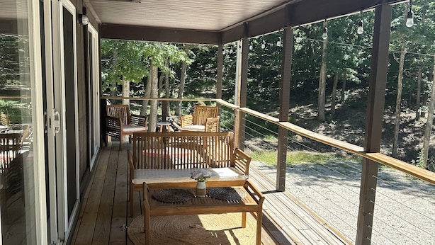 Una terraza de madera con zona de estar cubierta, que incluye una mesa y sillas, con vistas a un área boscosa.