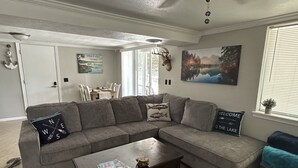 Una sala de estar con un sofá seccional gris, una mesa de centro y una alfombra. Hay un cuadro en la pared y una decoración con cabeza de ciervo.