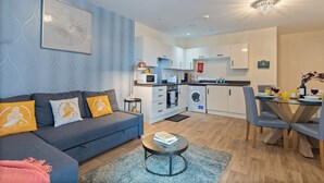 A modern apartment with a grey sofa, a dining table with chairs, a kitchen area, and a living room with a blue patterned wall and a framed artwork. - 2-BR serviced Apartment In Telford - W/Wi-fi /Netflix (Telford)