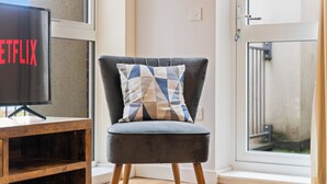 A modern living room with a dark grey armchair, a wooden TV stand, and a large window with a view of a balcony. - 2-BR serviced Apartment In Telford - W/Wi-fi /Netflix (Telford)
